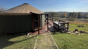 A yurt with a wooden deck leading to a covered area, surrounded by a grassy field and trees. - Luxury Yurt for Couples with Hot Tub on Deck (Glenwood)