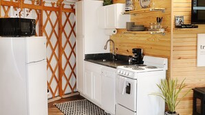 A compact kitchen with white cabinets, a stove, a sink, and a microwave. There is a wooden lattice partition, a small rug, and a sign that says "ENJOY." - Luxury Yurt for Couples with Hot Tub on Deck (Glenwood)