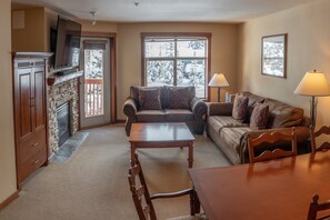 Cozy living room with a fireplace, sofa, dining table, and TV, offering a view of snow-covered trees.
