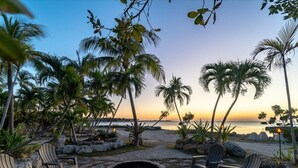 A beachside seating area with a fire pit, surrounded by palm trees and chairs, overlooking the ocean. - Sunsets in Paradise~ Gulf Front Pool Home, 22' Dock, Kayaks, Bikes & Paddleboards (Marathon)
