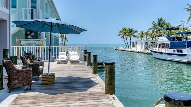 A wooden deck with wicker chairs and a blue umbrella, overlooking a marina with boats and palm trees. - Island Time~ Pool Home Seconds to Open Ocean! 26' Dock (Marathon)