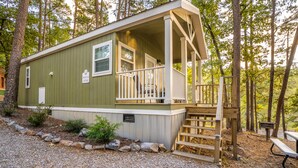 A small, single-story cabin with a porch and a deck, surrounded by trees.