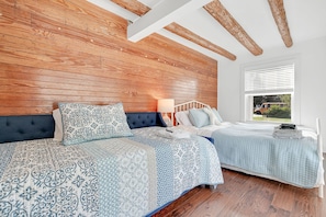 Une chambre avec deux lits, des poutres apparentes en bois au plafond, une grande fenêtre avec des stores et une vue sur l’extérieur.