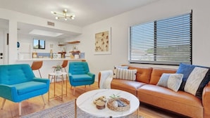 A modern living room with a blue sofa, a brown leather sofa, and a round coffee table. There is a kitchen area in the background with a window and blinds.
