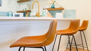 A kitchen area with a white countertop, a gold faucet, and three brown chairs with black metal legs.