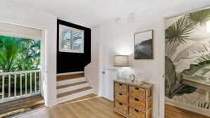 Una escalera de madera con escalones que conducen a una plataforma, una pared blanca con detalles negros, una mesita con una lámpara y una pared con un mural de plantas tropicales.