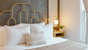 Una cama bien arreglada con cabecera dorada, una lámpara de noche y un espejo grande en la pared.