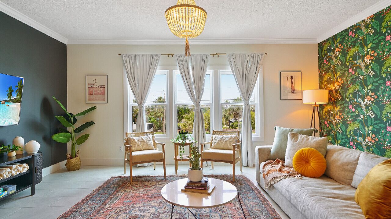 A living room with a round coffee table, two armchairs, a sofa, a chandelier, a large window with curtains, a plant, and a wall with a floral wallpaper.