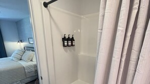Kylpyhuoneessa on lasiseinäinen suihkukaappi, wc ja pieni penkki. Viereisessä huoneessa näkyy sänky.