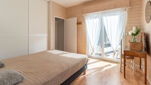 Ein Schlafzimmer mit einem Bett, Holzboden, einem Fenster mit Vorhängen, einem Stuhl und einem kleinen Tisch mit einem Korb.