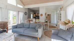 Ein geräumiges Wohnzimmer mit grauem Sofa, einem Couchtisch und einem Esstisch mit Stühlen. Im Hintergrund ist eine Küche zu sehen, und das Zimmer verfügt über große Fenster für natürliches Licht.
