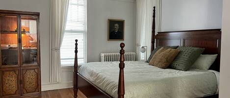 Une chambre à coucher avec un lit à baldaquin en bois, un radiateur, une armoire aux portes vitrées et un portrait accroché au mur.