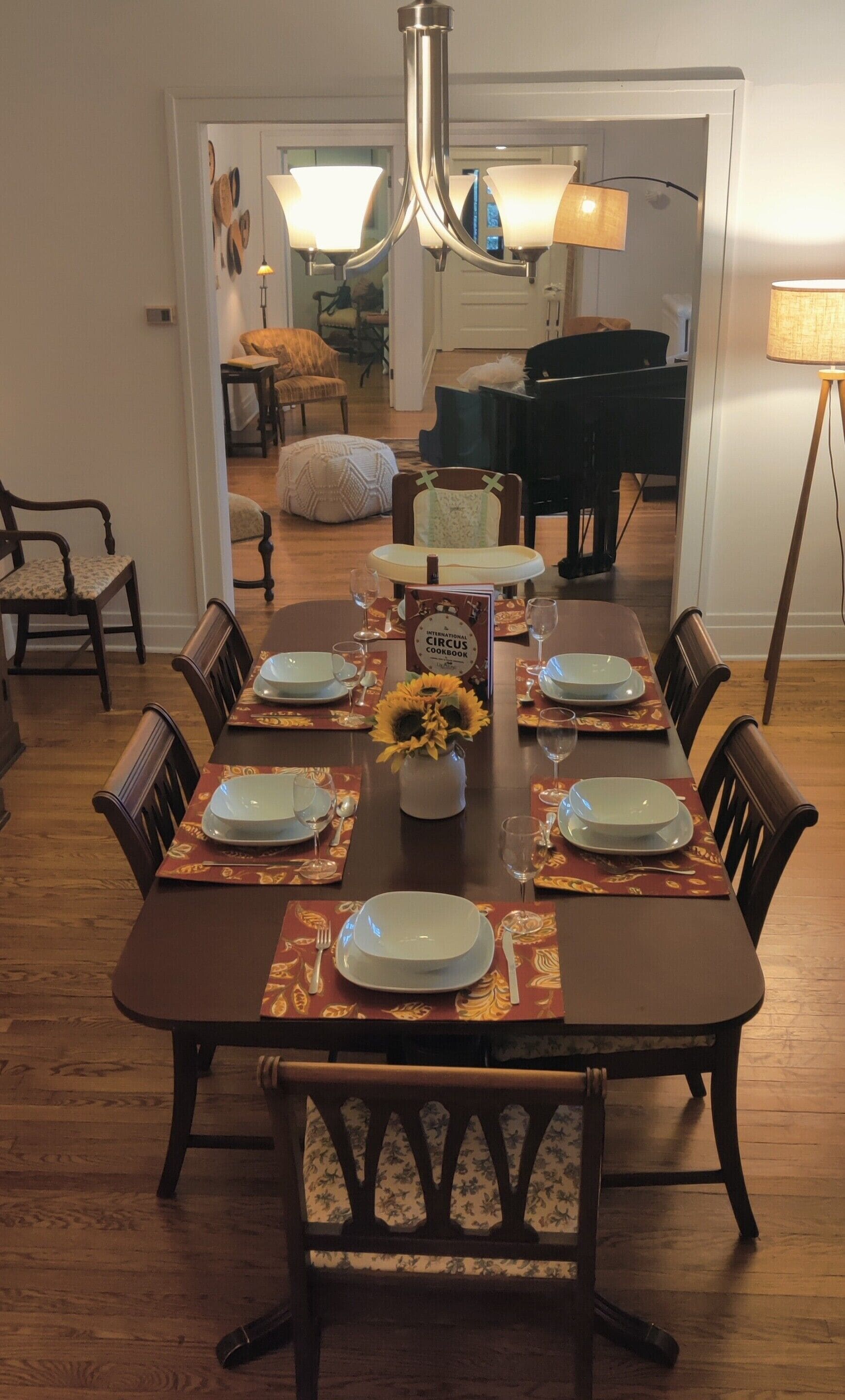 Un comedor con una mesa de madera dispuesta para seis personas, rodeada de sillas. Un libro titulado 'International Circus Cookbook' está sobre la mesa.