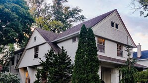 A two-story house with a steeply pitched roof, a chimney, and a well-maintained garden with various shrubs and a tall coniferous tree. - Historic Oak Park Home (Oak Park)