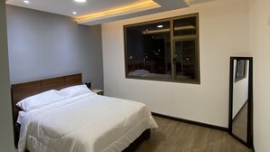 A modern bedroom with a large bed, recessed ceiling lighting, a window with a view of the city, and a wooden floor.