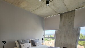 A modern bedroom with a large bed, a bedside table, a floor lamp, and a view of the city through large windows. - Loi Flats Neuquen, Edificio Talero (Neuquén)