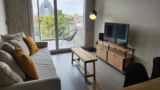 A modern living room with a sofa, a wooden coffee table, a TV on a wooden stand, and a balcony with chairs. - Loi Flats Neuquen, Edificio Talero (Neuquén)