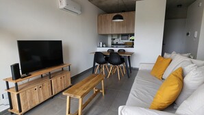 A modern living room with a flat-screen TV, wooden furniture, a sofa with yellow cushions, and a kitchen area in the background. - Loi Flats Neuquen, Edificio Talero (Neuquén)