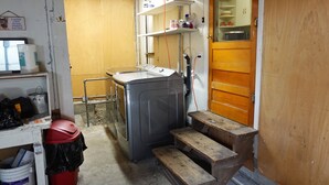 Un lavadero pequeño con lavadora, un estante con artículos de aseo, un banco de madera y una puerta que da a otra habitación.