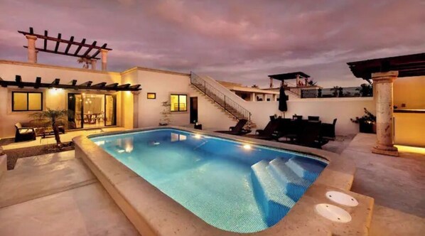 Relax by the clear blue pool surrounded by a well-maintained patio and pergola with soft lighting.
