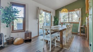 Un coin repas avec une table et des chaises en bois, une grande plante en pot et une vue sur l’extérieur grâce à une porte-fenêtre coulissante.