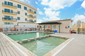 Un área de piscina en la azotea con piscina, sillones reclinables y un edificio con balcones.