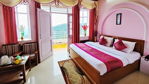 A hotel room with a bed, a desk, a chair, a window with curtains, and a view of the outdoors. - Hotel Madhuvan (Kandaghat)