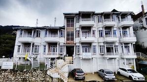 A multi-story hotel with a white facade, multiple balconies, and a parking area with several cars. - Hotel Madhuvan (Kandaghat)