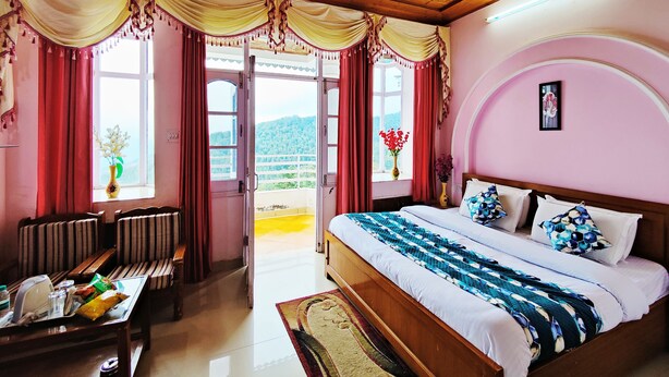 A bedroom with a bed, a desk, and a chair. There are curtains on the windows, and a ceiling fan is visible. - Hotel Madhuvan (Kandaghat)