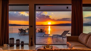 A room with a large sliding glass door leading to an outdoor area with a view of a sunset over water. Inside, there is a sofa and a small table with a decorative item.