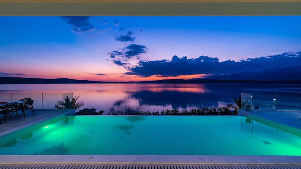 A pool with a view of a sunset over a calm lake, surrounded by chairs and plants.