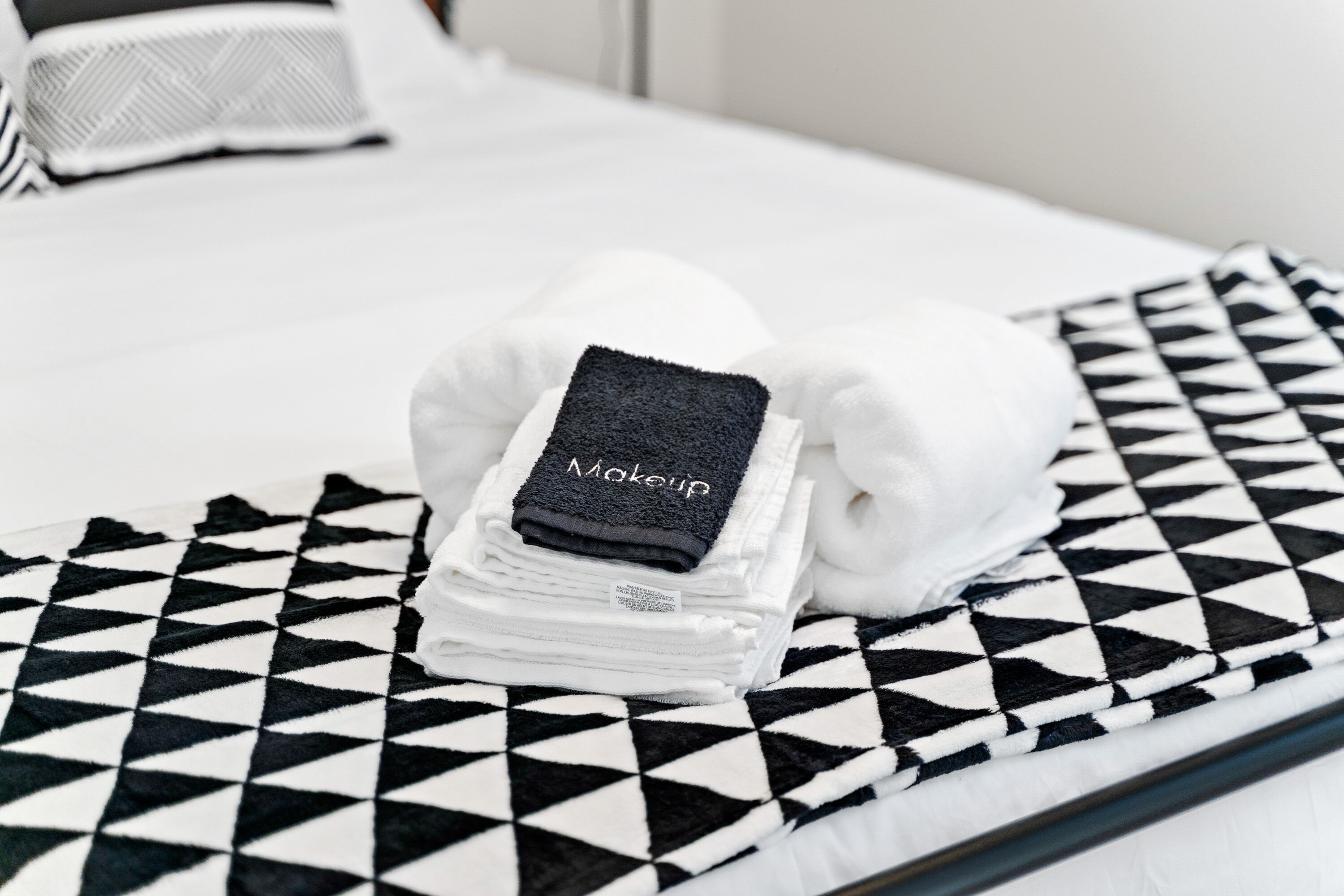 Una cama bien hecha con una funda de almohada a cuadros blanco y negro y una toalla doblada con la etiqueta 'Maquillaje'.