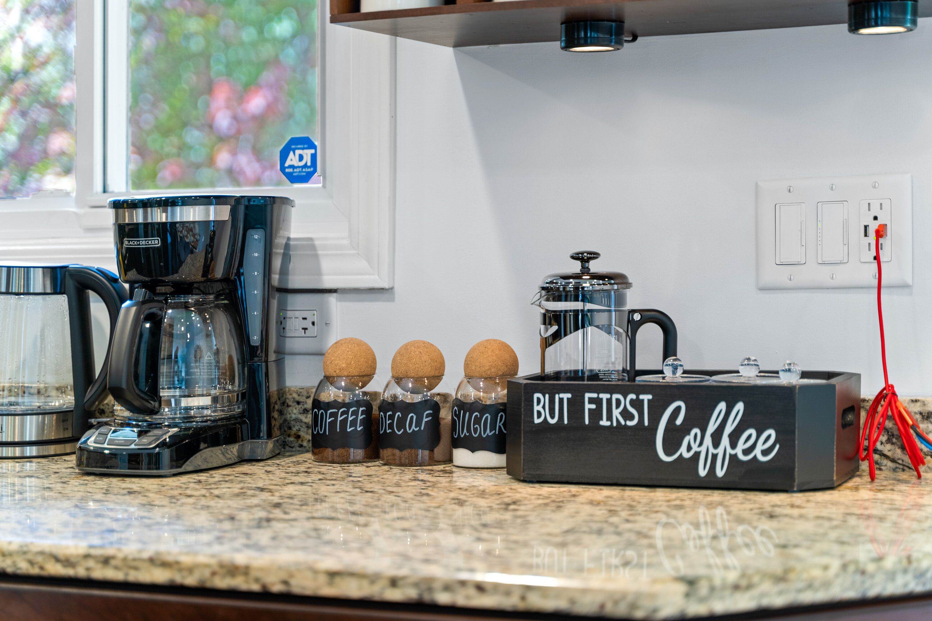 A kitchen counter with a coffee maker, sugar and decaf containers, and a coffee box labeled "But First Coffee".