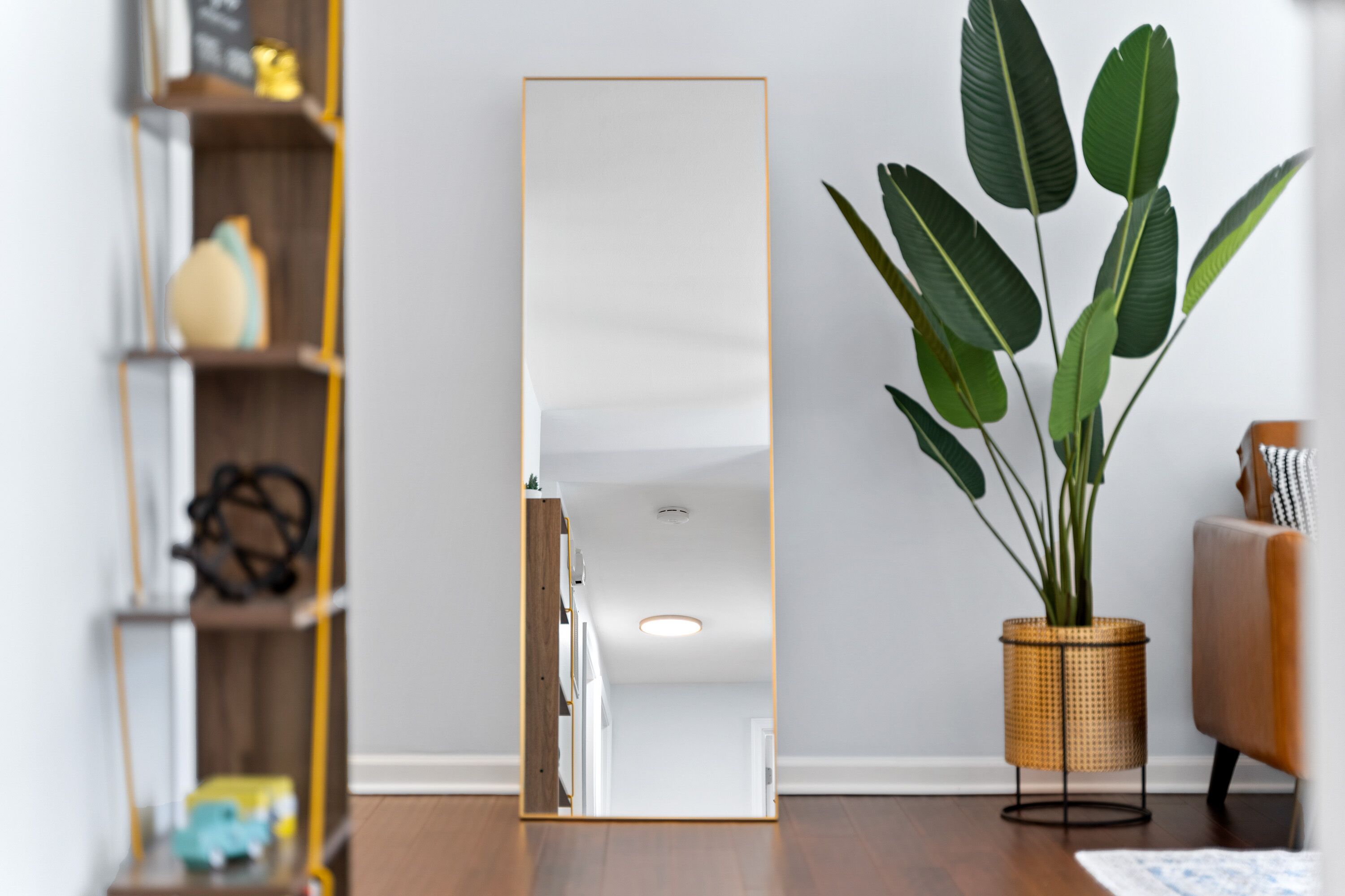 A room with a large mirror reflecting a hallway, a wooden floor, a wicker plant, and a bookshelf with various items.