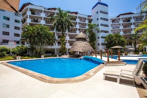 Ein Swimmingpool mit einer strohgedeckten Struktur, umgeben von Palmen und einem mehrstöckigen Gebäude mit Balkonen.