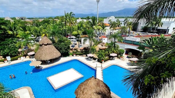A resort with a large swimming pool, thatched-roof structures, and palm trees. - CONDO MR BY TRVL2HM (Puerto Vallarta)