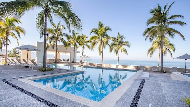 A pool area with lounge chairs and umbrellas, surrounded by palm trees, overlooking a beach.