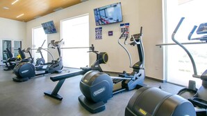 A gym with various exercise machines, a television mounted on the wall, and large windows allowing natural light.