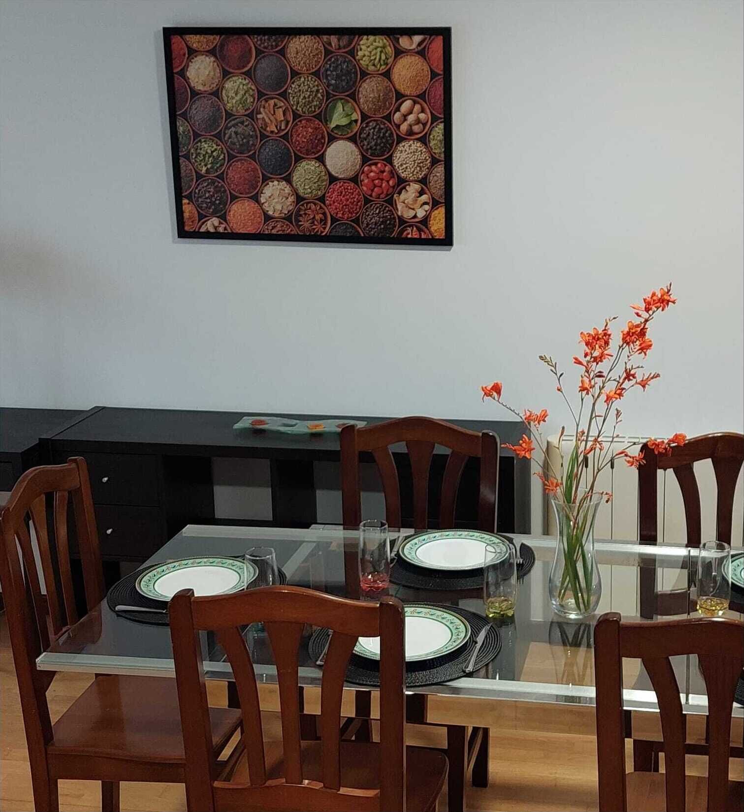 A dining area with a glass table, wooden chairs, a vase with orange flowers, and a wall art piece above the table.