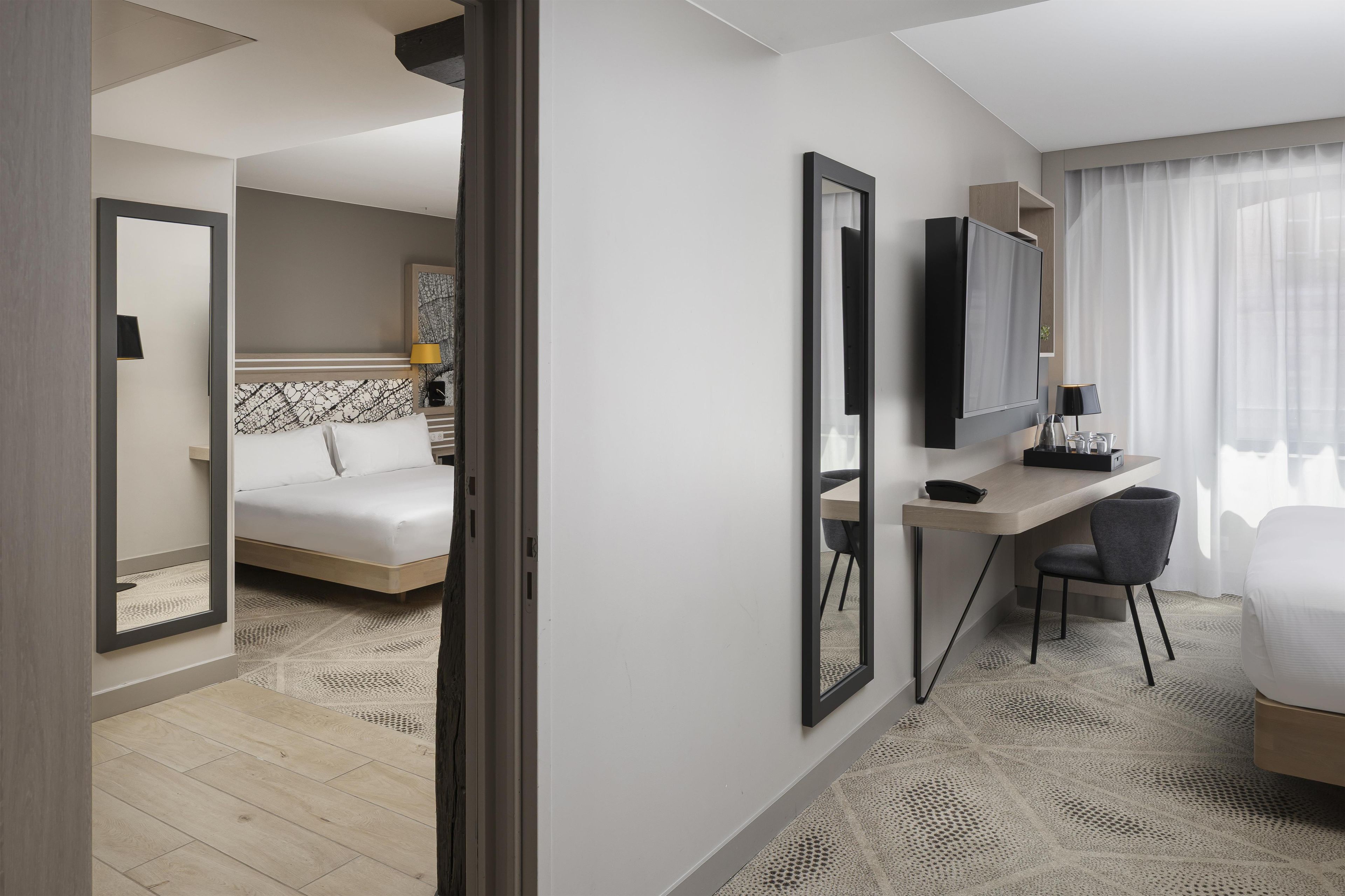 A hotel room with a bed, a desk, a chair, a TV, and a large mirror.