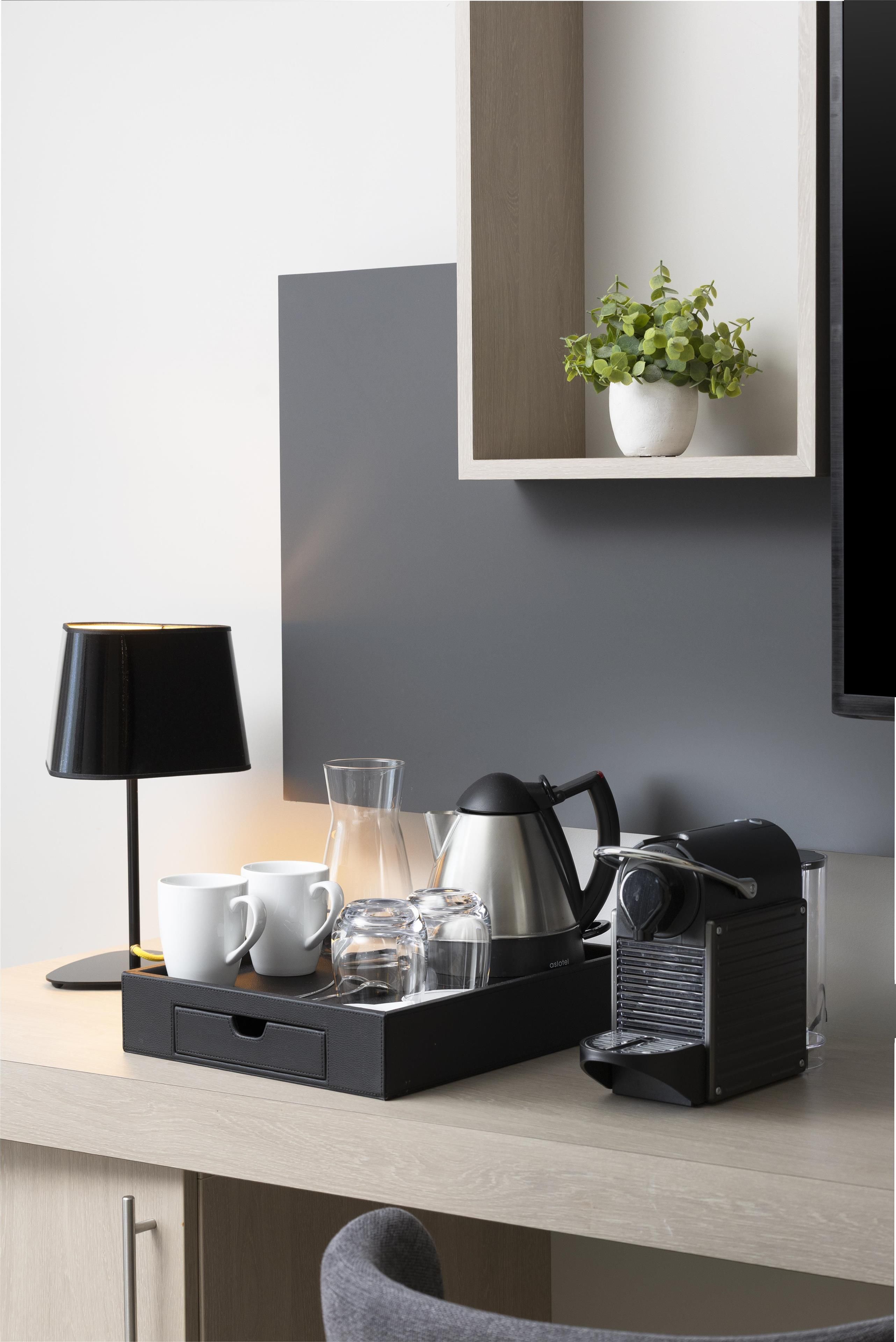 Ein moderner Küchenbereich mit Kaffeemaschine, Wasserkocher und Teekanne auf einem Tablett, einer Topfpflanze auf einer Ablage und einer Lampe auf der Arbeitsfläche.