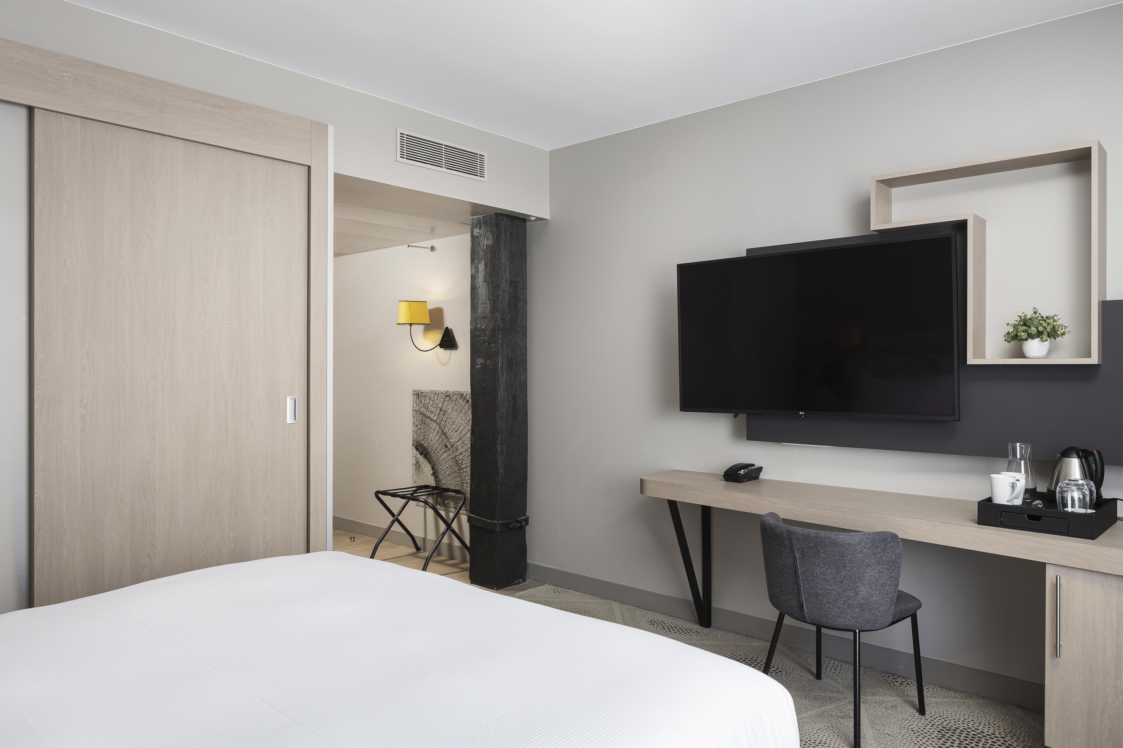 A hotel room with a bed, a desk with a chair, a TV, and a small shelf with a plant.