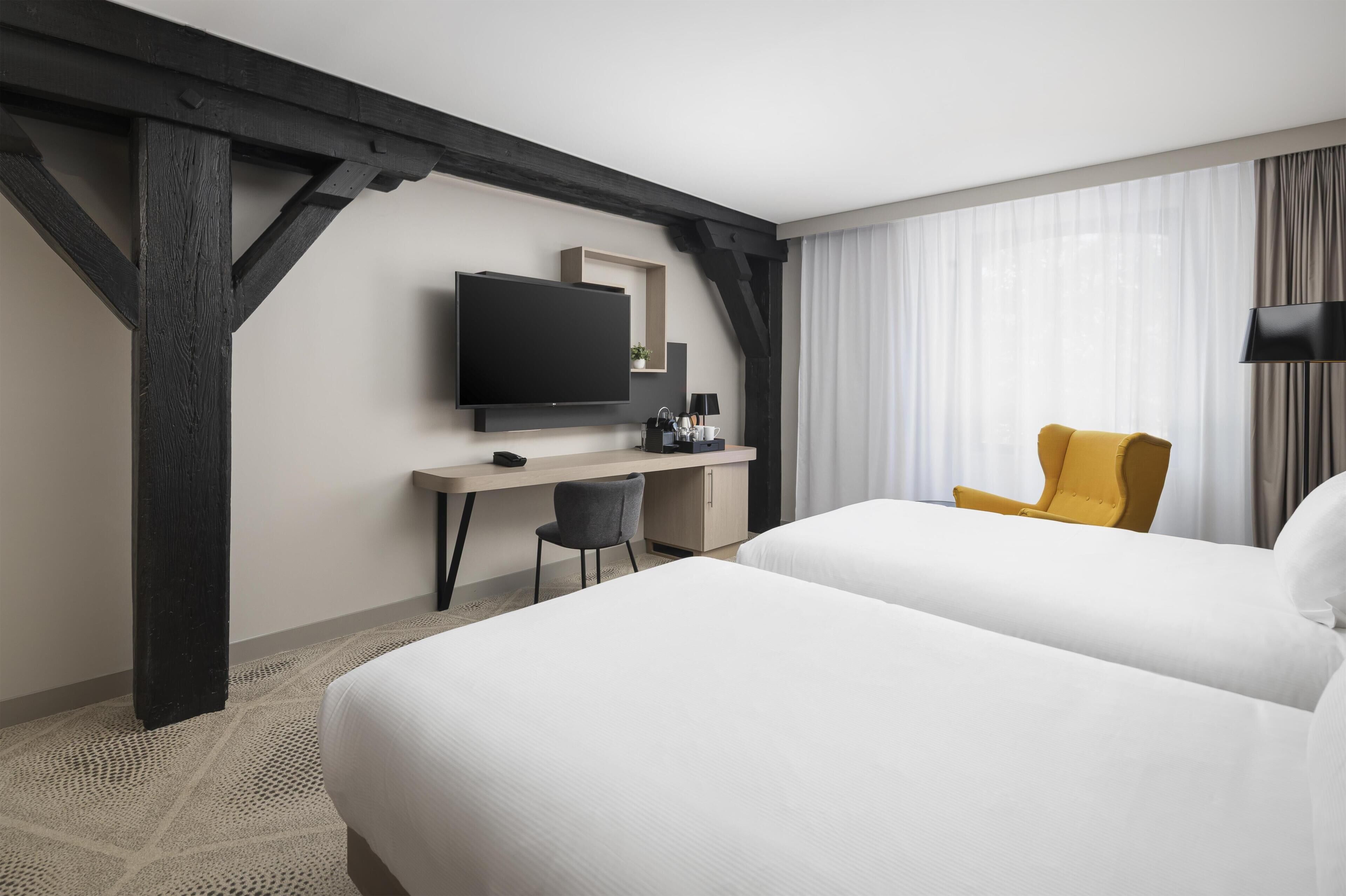 A hotel room with two beds, a desk with a TV, a chair, and a window with curtains.