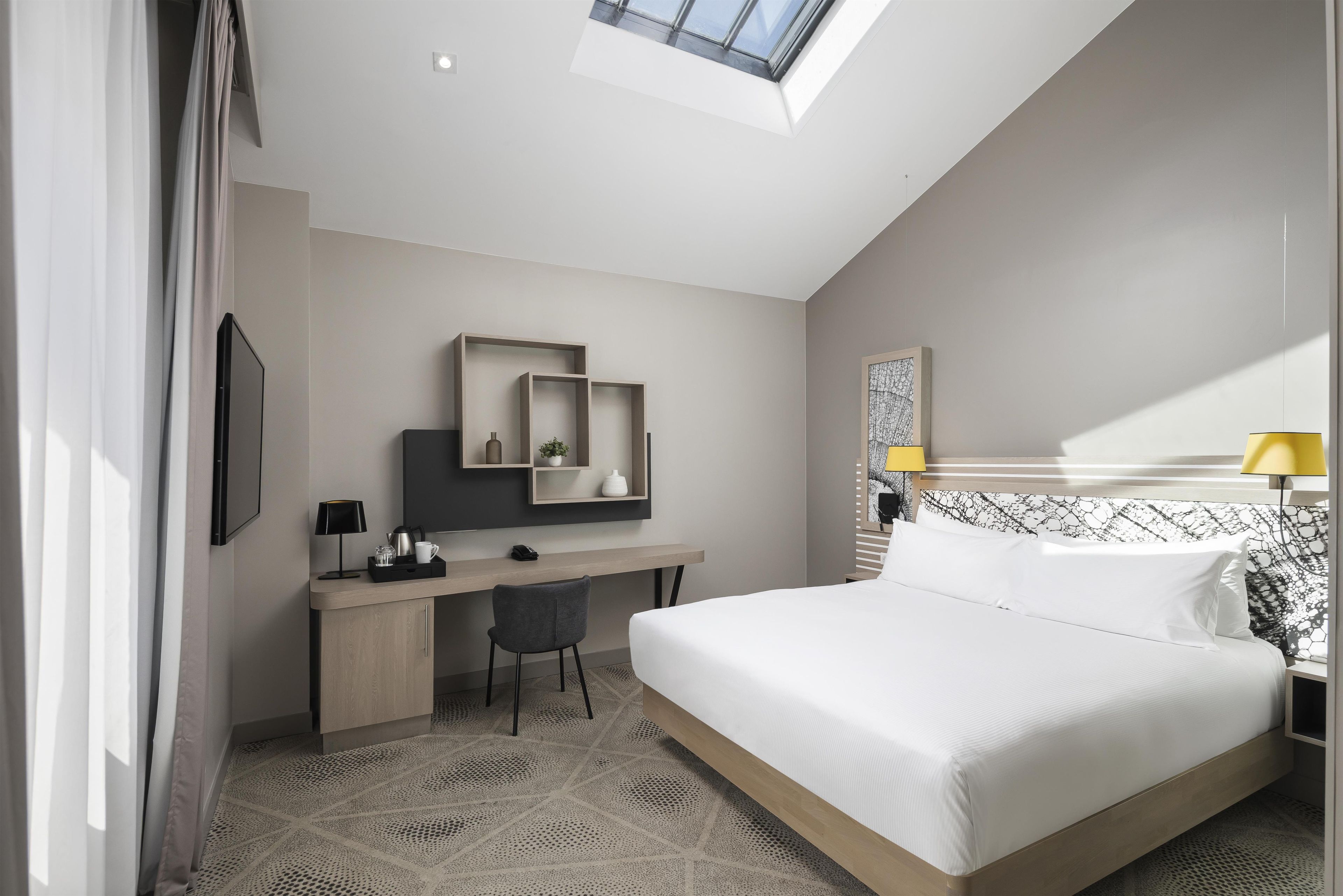 A modern hotel room with a skylight, a bed, a desk with a chair, and a bedside table with a lamp.