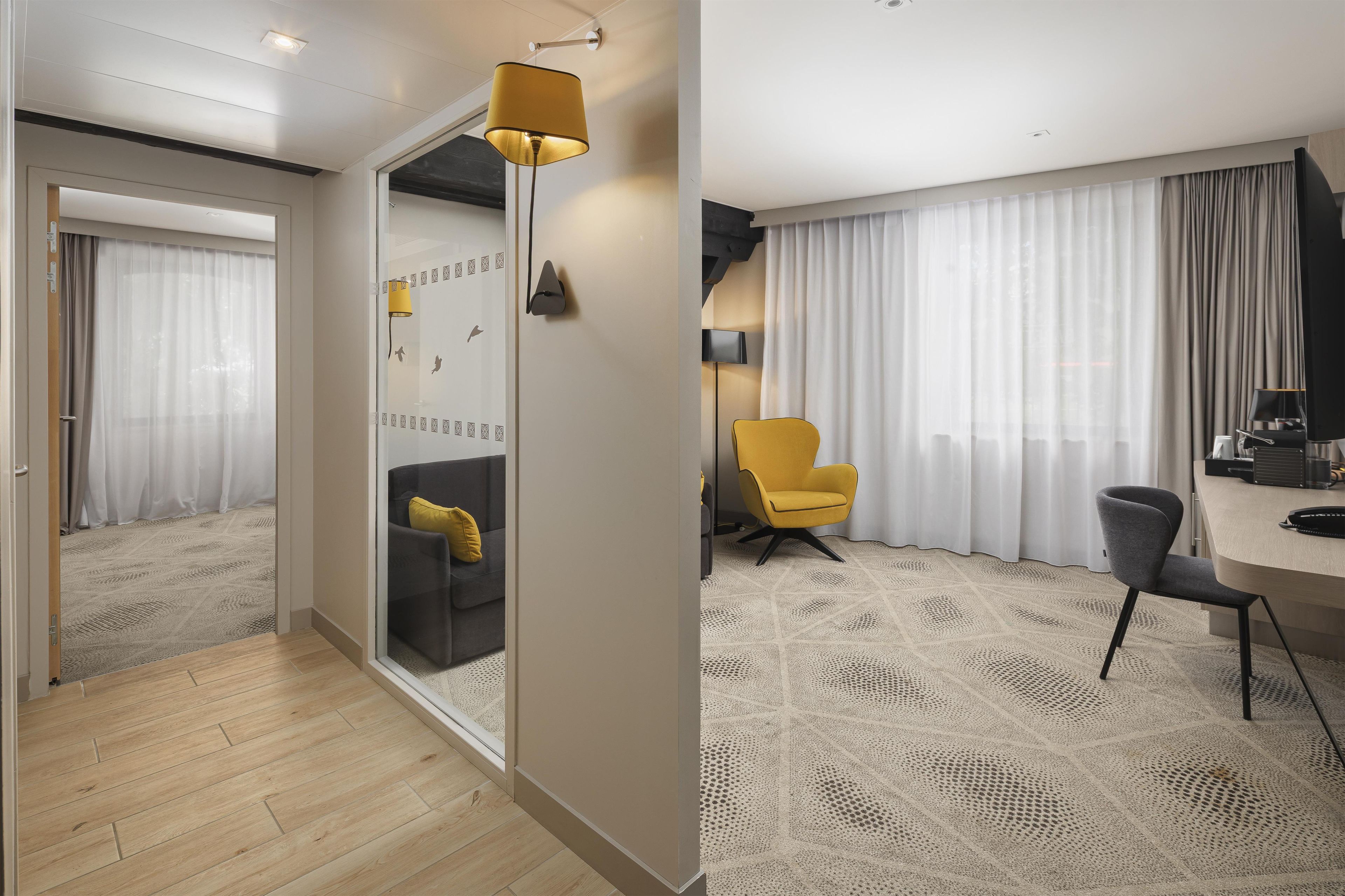A modern hotel room with a wooden floor, a glass partition, a yellow armchair, a desk with a chair, and a large window with curtains.