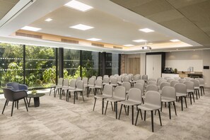 A conference room with rows of chairs, a projector, and a large window with a view of greenery. - Hilton Garden Inn Paris La Villette (Paris)