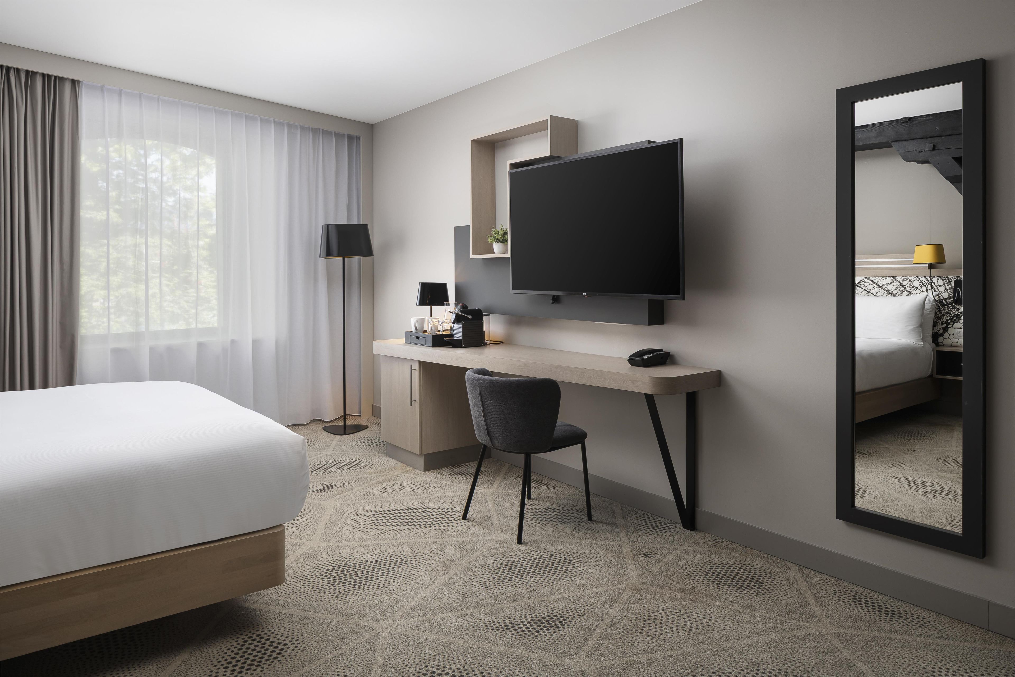 A hotel room with a bed, desk, chair, television, and a large mirror.