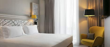 A hotel room with a large bed, a yellow armchair, a painting on the wall, and a window with curtains.