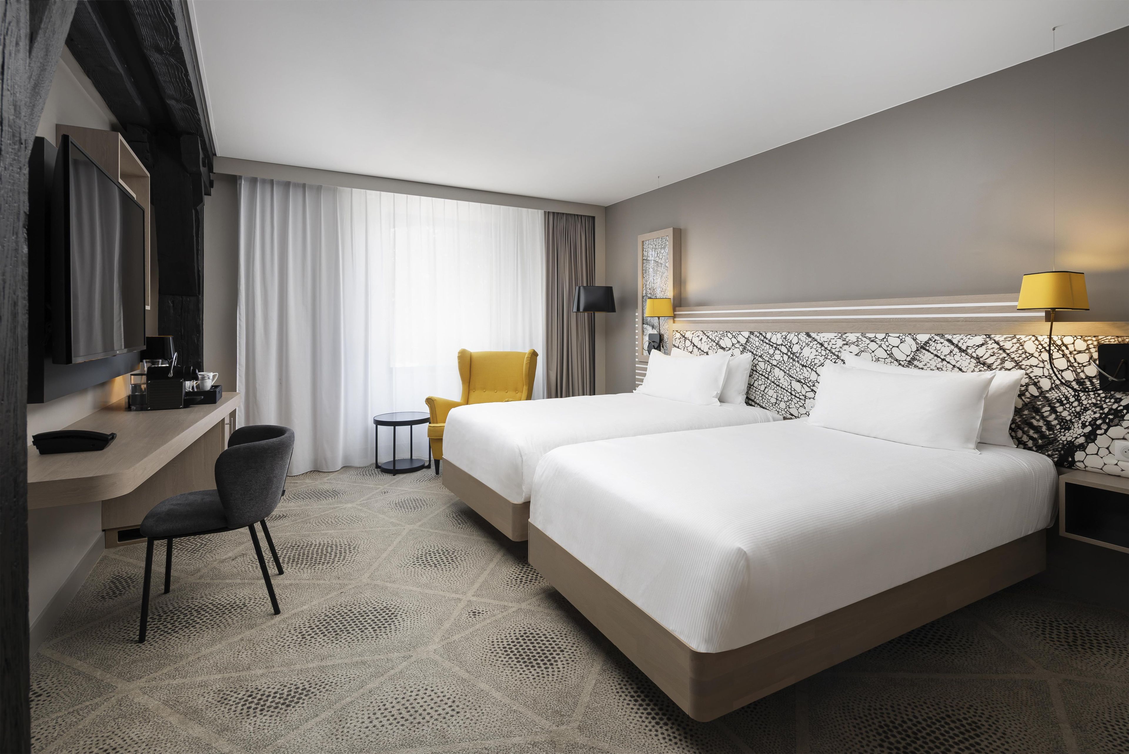Habitación de hotel con dos camas, un escritorio, una silla, una mesita y una lámpara.