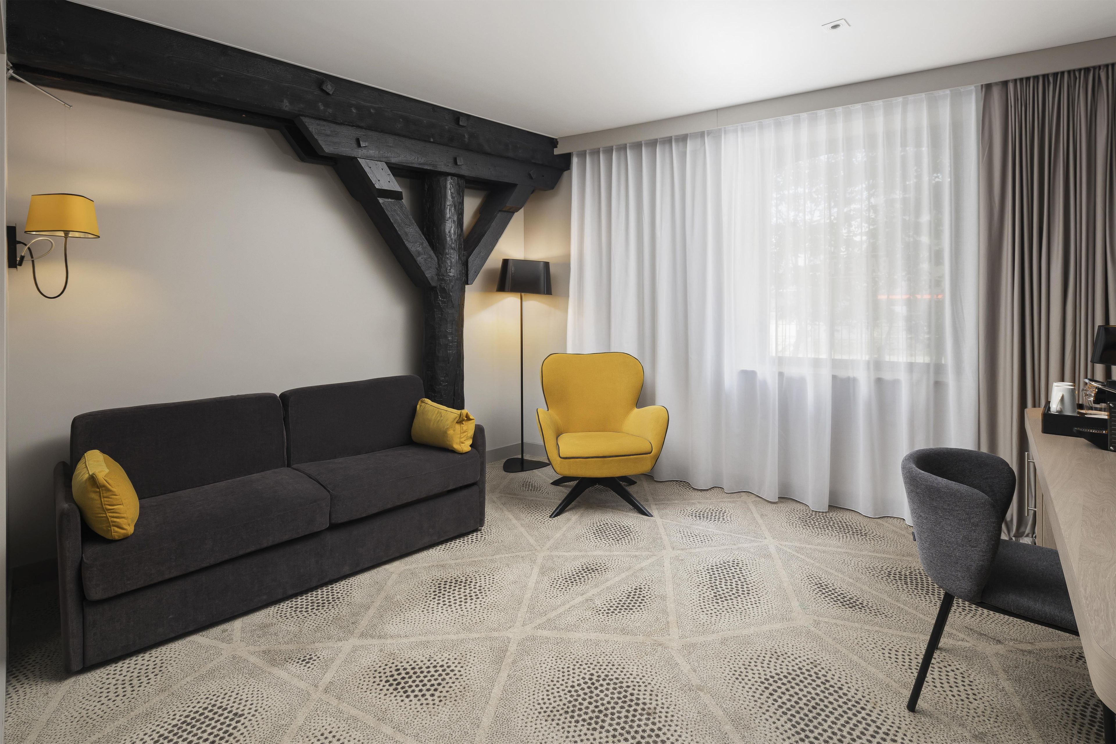 A modern hotel room with a dark grey sofa, a yellow armchair, and a small table with a lamp.